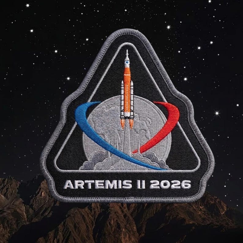 May include: A black and gray embroidered patch featuring the Artemis II 2026 mission logo. The design includes a rocket launching from a gray planet, with blue and red curved lines. The background shows a starry night sky and a brown mountain range.
