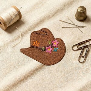 May include: A brown embroidered cowboy hat patch with pink, orange, and blue floral accents. The patch is surrounded by sewing supplies, including a spool of thread, needles, a thimble, and paper clips.