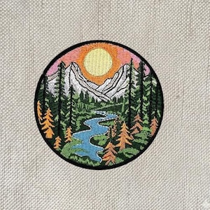 May include: Embroidered patch featuring a mountain landscape with a river, trees, and a setting sun. The patch has a black border and is circular. The colors include orange, pink, green, blue, and white.