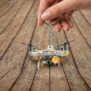 May include: A white drone keychain with the name "ALEX" printed on it. The keychain is decorated with small yellow lights and is suspended from a silver keyring and chain. The background is a wooden surface.