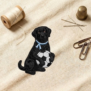 May include: An embroidered patch featuring a black Labrador retriever dog with a light blue collar, sitting and holding a black and white soccer ball. The patch is surrounded by a wooden spool of thread, a metal thimble, needles, and paper clips.