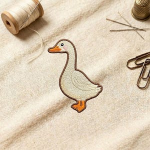 May include: An embroidered patch of a goose with a brown outline, orange beak and feet, and a pink cheek. The patch is on a beige fabric, with sewing supplies including thread, needles, and a thimble in the background. The goose is facing left.