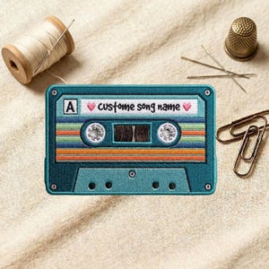 May include: Embroidered teal cassette tape patch with the text "custom song name" and a heart symbol. The patch has a retro design with colorful stripes. A spool of thread, needles, a thimble, and paper clips are also in the image.