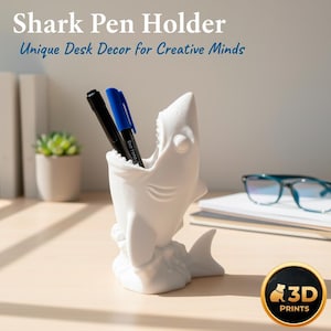 May include: A white shark pen holder with a wide open mouth, holding a black and blue pen. The text "Shark Pen Holder" and "Unique Desk Decor for Creative Minds" are at the top. The pen holder is on a light brown desk.
