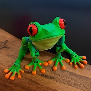 May include: A vibrant green frog figurine with large red eyes and orange-tipped feet. The frog is posed on a wooden surface, showcasing its articulated limbs and detailed features. The toy frog is a playful representation of a tree frog.