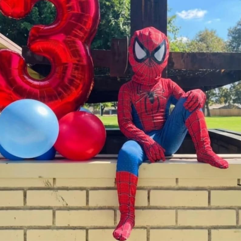 May include: A child dressed in a Spider-Man costume, sitting on a brick wall. The costume is red and blue with a spider emblem on the chest. A large red number 3 balloon and two smaller balloons, one red and one blue, are next to the child.
