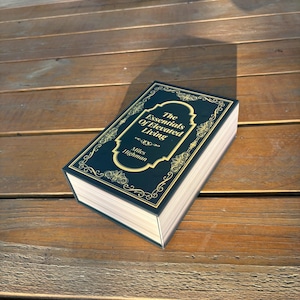 May include: A hardcover book titled "The Essentials of Elevated Living" by Miles Highman. The book has a dark teal cover with gold lettering and decorative accents. The book is resting on a wooden surface.