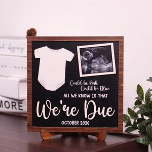 May include: Wooden framed pregnancy announcement sign with a black background. Features a white onesie graphic, ultrasound photo, and the text "Could be Pink, Could be Blue, All We Know Is That We're Due October 2026."