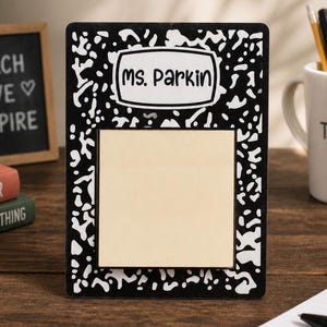 May include: A black and white composition notebook-style picture frame with the name "Ms. Parkin" on it. The frame has a blank, light-colored center. A mug that says "Best Teacher Ever" is in the background.