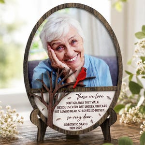 May include: An oval wooden memorial plaque featuring a photograph of an older person, a small red cardinal figurine perched on a dark wooden branch, and text that reads: Those we love don't go away they walk beside us every day. Unseen, unheard, but always near. So loved, so missed. So very dear. Dorothy C. Sarto 1928-2025.