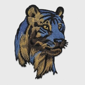 May include: An embroidered patch featuring the head of a tiger in profile, rendered in shades of metallic gold, deep blue, and black thread. The design captures the feline's facial features, including its eyes, ears, and whiskers in detail.