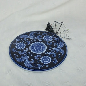 May include: A round, dark blue ceramic coaster with a floral design in shades of blue and white. The coaster features a detailed pattern of flowers and leaves. A decorative metal spider web is next to the coaster.