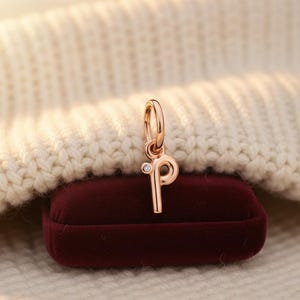 May include: A rose gold charm shaped like the letter "P", featuring a small diamond. The charm hangs from a ring and sits on a burgundy velvet box, with a cream-coloured knitted backdrop.