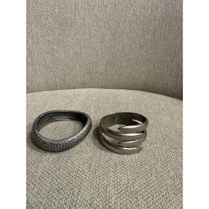 May include: Two silver-toned metal bracelets. One bracelet features a textured, diamond-patterned surface with a hinged closure. The second bracelet has a smooth, multi-layered wrap design with pointed ends and a hinged mechanism for opening.