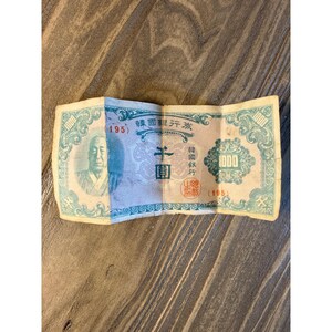 May include: A vintage 1000 won banknote featuring a portrait of a person on the left, intricate teal and cream decorative borders, the number 1000 in the corners, and red stamps. The paper currency displays Korean text and the number 195.