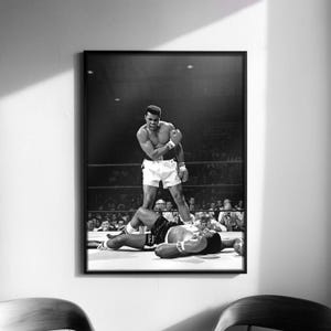 Puede incluir: Fotografía enmarcada en blanco y negro de un combate de boxeo. Un boxeador se alza sobre su oponente derrotado en el ring. La imagen captura un momento de victoria y derrota, con una multitud en el fondo.