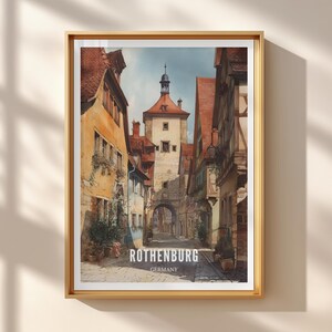 Könnte beinhalten: Ein gerahmter Druck mit einem Aquarellgemälde von Rothenburg, Deutschland. Das Kunstwerk zeigt eine schmale Straße mit farbenfrohen Gebäuden, einem zentralen Turm und dem Text "ROTHENBURG GERMANY" am unteren Rand.