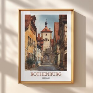 Può includere: Una stampa incorniciata con un dipinto ad acquerello di Rothenburg, in Germania. L'opera raffigura una strada stretta fiancheggiata da edifici colorati e tetti di tegole rosse, che conduce a un'alta torre. La stampa è in una cornice dorata, con la scritta "ROTHENBURG" e "GERMANY" in basso.