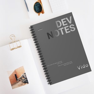 May include: A gray spiral-bound notebook with the text "DEV NOTES" in silver. The notebook also has the year "2020" printed on it, along with the word "Vido" and a quote. A gold paper clip and sunglasses are also visible.
