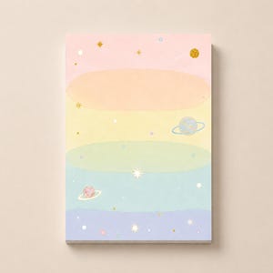 May include: A rectangular notepad featuring a horizontal gradient background of pastel pink, orange, yellow, green, blue, and purple. The design is decorated with small gold stars, white dots, and illustrations of three planets with rings.