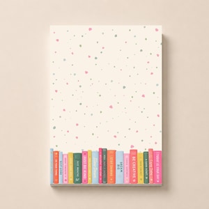 May include: A rectangular notepad with a cream-colored background featuring a pattern of small pink and green dots. At the bottom, a row of colorful book spines is illustrated, each labeled with motivational phrases like Dream Big and Be Creative.