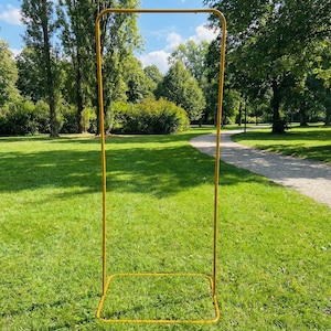 May include: A yellow metal archway stands on a grassy lawn in a park setting. The archway is rectangular, with a square base and a top frame. The background features trees, a walking path, and a blue sky.