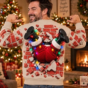 May include: A beige long-sleeve jumper featuring a 3D graphic of Santa Claus's legs protruding from a brick wall design. The jumper includes a string of colourful LED lights wrapped around the legs and a textured brick pattern on the back.