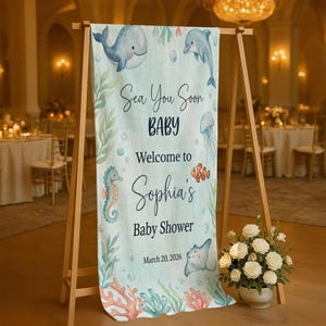 May include: A vertical light blue banner on a wooden stand featuring illustrations of a whale, dolphin, jellyfish, seahorse, and stingray. The text reads: Sea You Soon BABY Welcome to Sophia's Baby Shower March 20, 2026. Coral and seaweed accents.