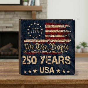 Op de afbeelding: Vierkante houten onderzetter met een verweerd ontwerp van de Amerikaanse vlag. De onderzetter heeft de tekst "1776", "We The People", "250 YEARS" en "USA" met steraccenten. Het kleurenschema omvat rood, wit en blauw.