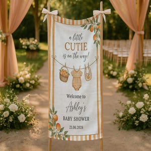 May include: A vertical fabric banner featuring orange and white stripes, decorated with illustrations of oranges, a basket, a baby onesie, and a bib. The text reads: a little CUTIE is on the way! Welcome to Ashley's BABY SHOWER 21.06.2026.