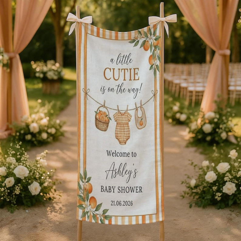 May include: A vertical fabric banner featuring orange and white stripes, decorated with illustrations of oranges, a basket, a baby onesie, and a bib. The text reads: a little CUTIE is on the way! Welcome to Ashley's BABY SHOWER 21.06.2026.