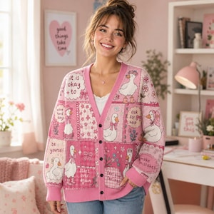 Op de afbeelding: Een roze vest met V-hals voorzien van een patchworkontwerp met witte eenden, bloemmotieven en teksten waaronder "it is okay to", "have hard days", "be yourself", "ask for help", "make mistakes", "start over again" en "feel your feelings".