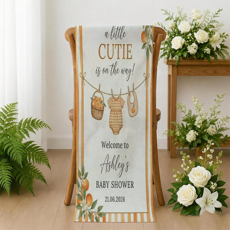 May include: A vertical rectangular banner featuring the text 'a little CUTIE is on the way!' and 'Welcome to Ashley's BABY SHOWER 21.06.2026'. The design includes illustrations of a basket of oranges, a checkered baby onesie, and a bib on a clothesline.