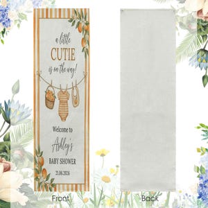 May include: A rectangular fabric banner featuring orange stripes on the edges and a central design with the text a little CUTIE is on the way! Welcome to Ashley's BABY SHOWER 21.06.2026, illustrated with a clothesline, basket, onesie, and bib.