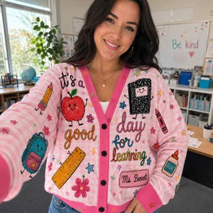 Puede incluir: Un cárdigan de cuadros vichy rosa claro y blanco con gráficos de temática escolar que incluyen una manzana, un lápiz, una regla, una mochila, un bote de pegamento y un cuaderno. El texto dice it's a good day for learning y Ms. Bennett.