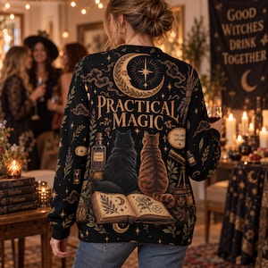 May include: A black long-sleeve sweatshirt featuring a graphic print on the back with the text "PRACTICAL MAGIC," a crescent moon, an open book, a black cat, a tabby cat, potion bottles, and botanical illustrations in gold and brown tones.