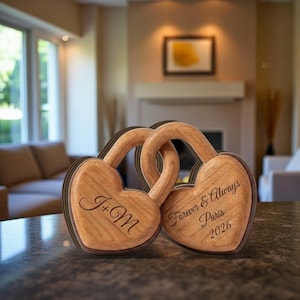 May include: Two wooden heart-shaped lock sculptures with interlocking handles. One heart is engraved with "J+M", the other with "Forever & Always Paris 2026". The wood has a light brown color with darker brown edges.
