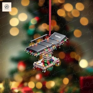 May include: A miniature, black surgical table Christmas ornament, adorned with colorful string lights. The ornament has the word "NAME" printed on it. The ornament is hanging from a red ribbon against a blurred background of Christmas lights.