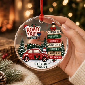 May include: A round, clear acrylic Christmas ornament featuring a red car with a wreath and a snowman, a road trip signpost with city names, and the text ROAD TRIP 2026 and SHAFER FAMILY VACATION. The ornament hangs from a red ribbon.
