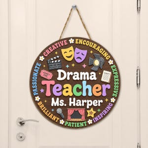 May include: A round wooden hanging sign featuring the text Drama Teacher Ms. Harper in the center. The design includes colorful icons of theater masks, a clapperboard, a spotlight, a script, a microphone, and a stage with curtains, surrounded by words.