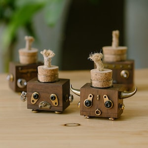 May include: Wooden bull-shaped bottle stoppers with brass accents and cork tops. The stoppers have a rustic, steampunk aesthetic, with small metal details and a natural wood finish. The horns are gold-colored.