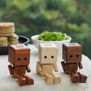 Può includere: Tre figurine di robot in legno in varie tonalità di marrone e beige. Ogni robot ha una testa a forma di scatola con due linee sulla parte superiore e occhi circolari. I robot hanno braccia e gambe articolate.
