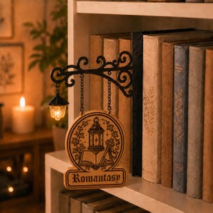 May include: A black metal bookend featuring a decorative scrollwork arm that holds a miniature glowing lantern and a circular wooden sign. The sign displays an engraved illustration of a lantern, an open book, and a key, with the text Romantasy.