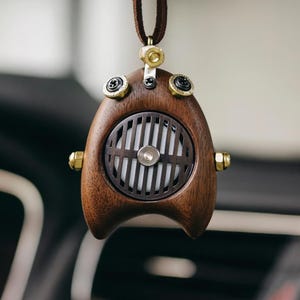 Puede incluir: Un ambientador de madera con un diseño único, con un acabado marrón oscuro y detalles metálicos. El ambientador tiene una rejilla circular en el centro y está suspendido por un cordón de cuero marrón. El diseño incorpora tornillos y pernos de color latón.