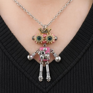 Könnte beinhalten: Eine silberfarbene Halskette mit einem Roboteranhänger. Der Roboter hat einen rosa Körper, grüne Augen und eine goldene Krone. Die Arme und Beine des Roboters sind silberfarben, mit kugelförmigen Händen und Füßen. Die Halskette wird vor einem schwarzen Hintergrund getragen.