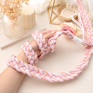 Puede incluir: Cuerda trenzada y pulseras en tonos rosa, blanco y melocotón. La cuerda es gruesa y parece estar hecha de tela suave. Las pulseras tienen un diseño a juego. Los artículos se exhiben sobre una superficie clara, con elementos decorativos en el fondo.