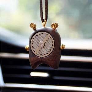 Puede incluir: Un ambientador para coche de madera marrón con un diseño retro, con una rejilla metálica circular y detalles dorados. Cuelga de un cordón de cuero marrón, añadiendo un toque vintage al interior del coche.