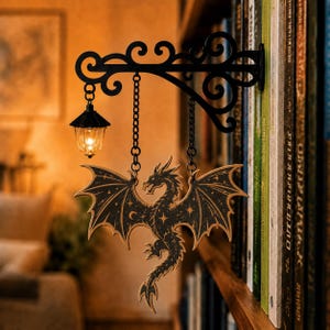 May include: A black metal wall-mounted bracket with a decorative scroll design, featuring a small hanging lantern and a flat, laser-cut wooden dragon ornament decorated with gold-colored celestial stars and moon patterns suspended by chains.