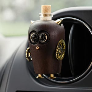 Può includere: Un diffusore per auto a forma di gufo marrone scuro con dettagli color oro. Il gufo ha un tappo di sughero, dettagli laterali a forma di ingranaggio e piedini color oro. Gli occhi sono neri con una superficie riflettente e le orecchie sono punte dorate.