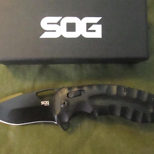 May include: A black SOG folding knife with a curved blade and textured handle, positioned below a black box with the white SOG logo. The knife has a partially serrated edge and a gold-colored blade tip.